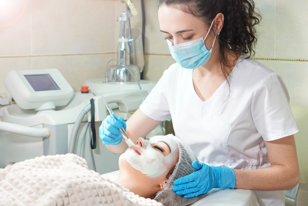 iStock 1206317911 1024x688 - Cosmetologist doing cosmetic face mask treatment for the young woman. -